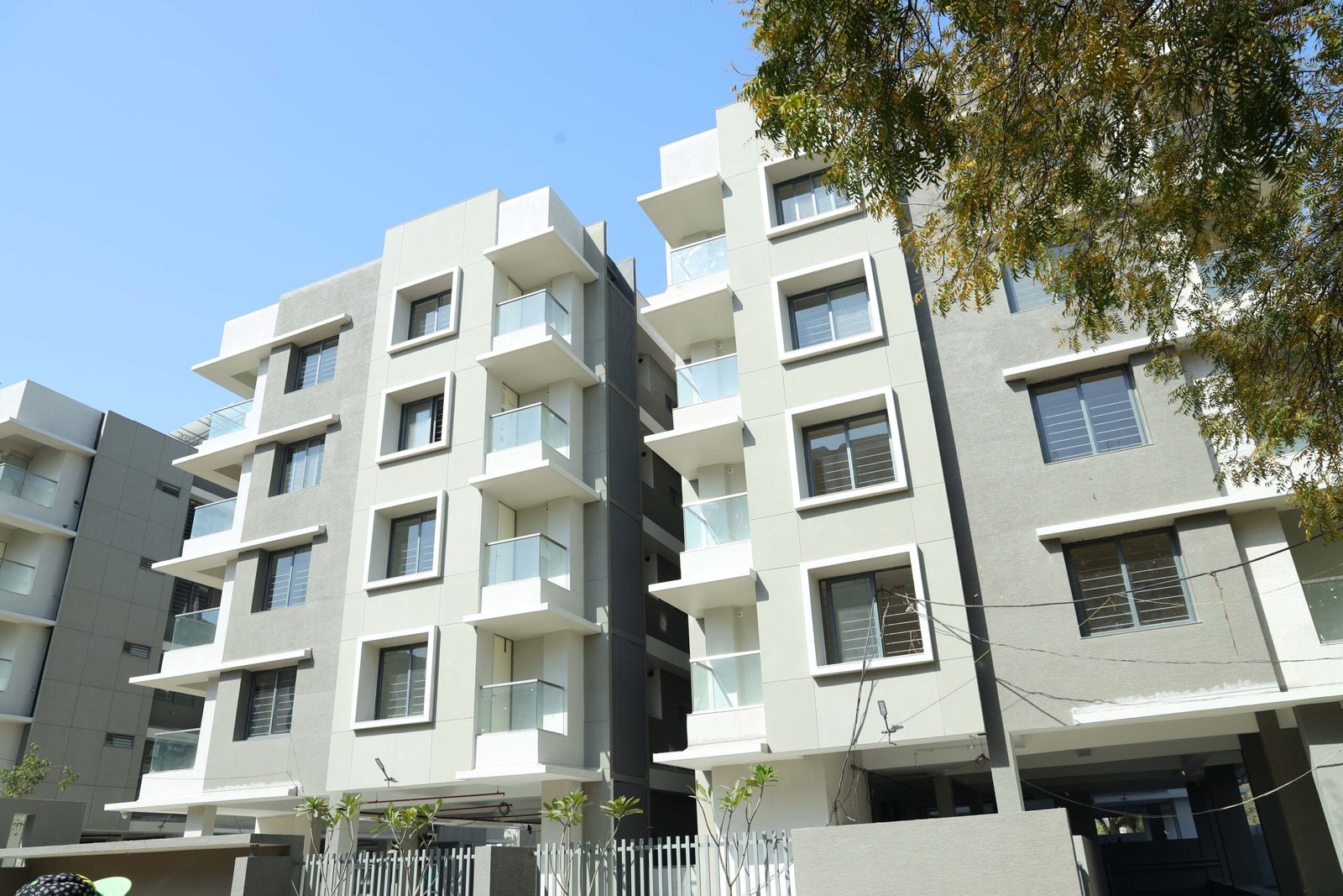 - Front elevation of Arise Ample low-rise luxury apartments on S.G. Highway, Ahmedabad - Solar panels and energy-efficient fixtures integrated on the rooftop of Arise Ample residences - Dramatic fenestration pattern across the monotone façade of Arise Ample apartments - Achromatic color scheme with bold planes and shaded tints on the Arise Ample exterior - Landscaped courtyard promoting healthy living at the environmentally friendly Arise Ample complex