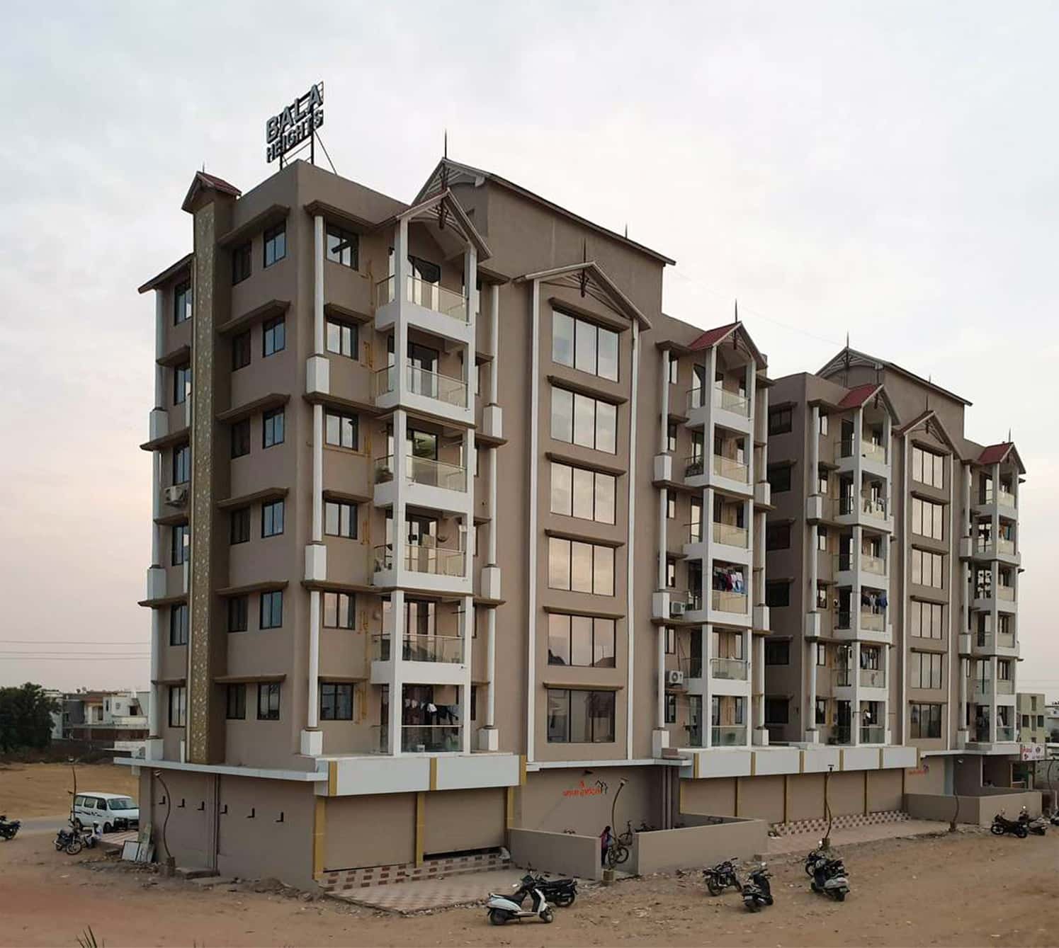 - Front elevation of Bala Heights high-rise in Mehsana, showcasing traditional motifs and modern glass accents - Ornate wrought-iron balcony details on the classic facade of Bala Heights residential tower - Street-level view of ground-floor shops integrated into the Bala Heights mixed-use development - Interior perspective of a 2 BHK apartment at Bala Heights, blending heritage details with contemporary finishes - Landscaped communal courtyard at Bala Heights, featuring greenery and seating areas for residents