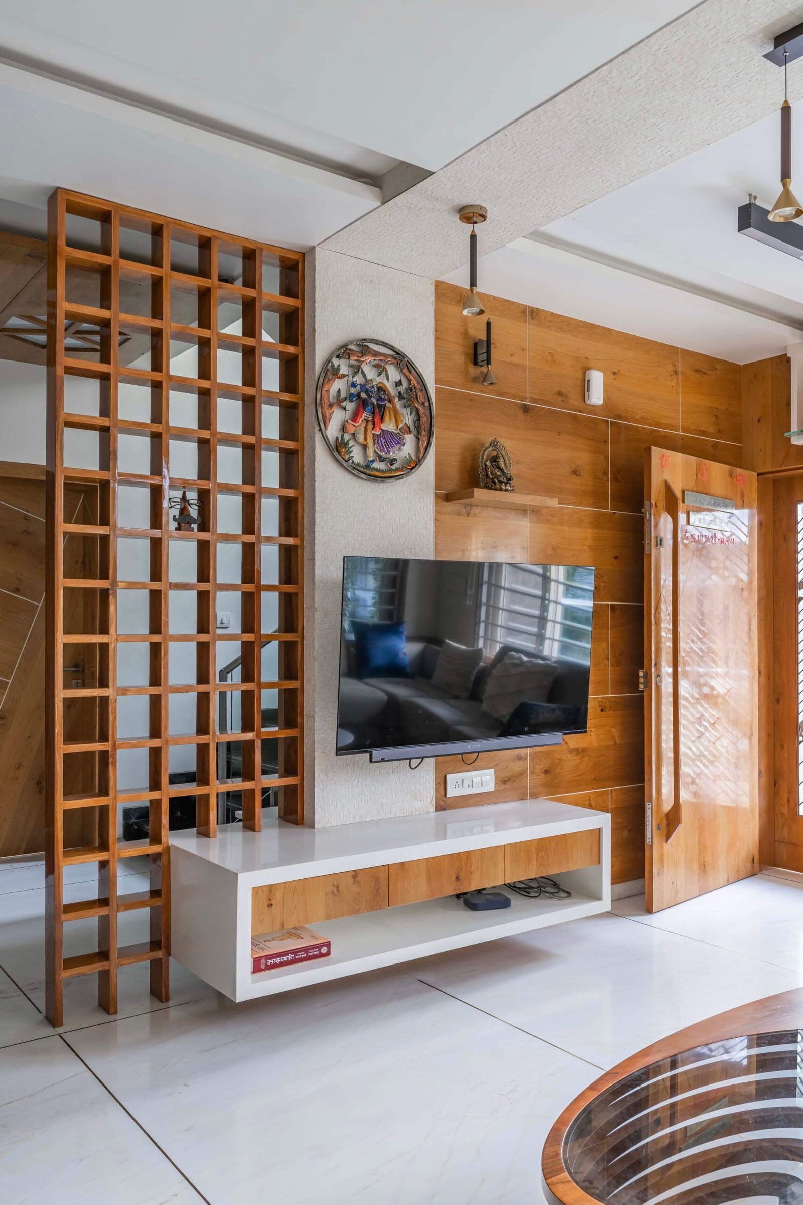 - Modern bungalow facade with exposed concrete, stone cladding, and wooden louvers in Ahmedabad - Sandstone-clad entrance with illuminated staircase and landscaped exterior - Open-plan living and dining area with geometric partition and modern chandelier - Master bedroom with blue textured wallpaper, wooden ceiling grid, and floating bedside units - Guest bedroom with geometric headboard, floating desk, and accent lighting - Polished flooring, natural wood finishes, and integrated lighting in modern residential interior