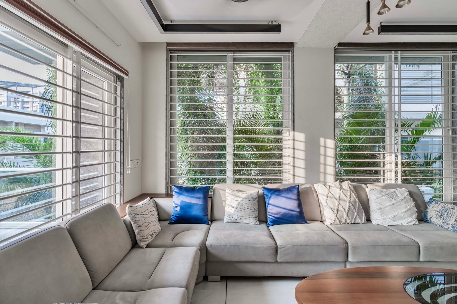 - Modern bungalow facade with exposed concrete, stone cladding, and wooden louvers in Ahmedabad - Sandstone-clad entrance with illuminated staircase and landscaped exterior - Open-plan living and dining area with geometric partition and modern chandelier - Master bedroom with blue textured wallpaper, wooden ceiling grid, and floating bedside units - Guest bedroom with geometric headboard, floating desk, and accent lighting - Polished flooring, natural wood finishes, and integrated lighting in modern residential interior