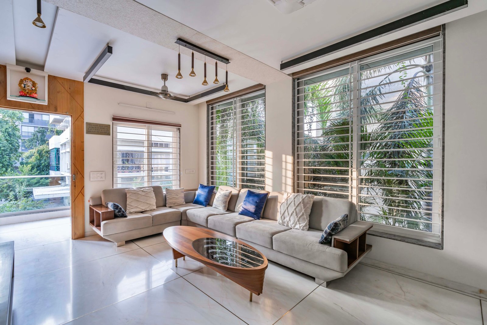 - Modern bungalow facade with exposed concrete, stone cladding, and wooden louvers in Ahmedabad - Sandstone-clad entrance with illuminated staircase and landscaped exterior - Open-plan living and dining area with geometric partition and modern chandelier - Master bedroom with blue textured wallpaper, wooden ceiling grid, and floating bedside units - Guest bedroom with geometric headboard, floating desk, and accent lighting - Polished flooring, natural wood finishes, and integrated lighting in modern residential interior