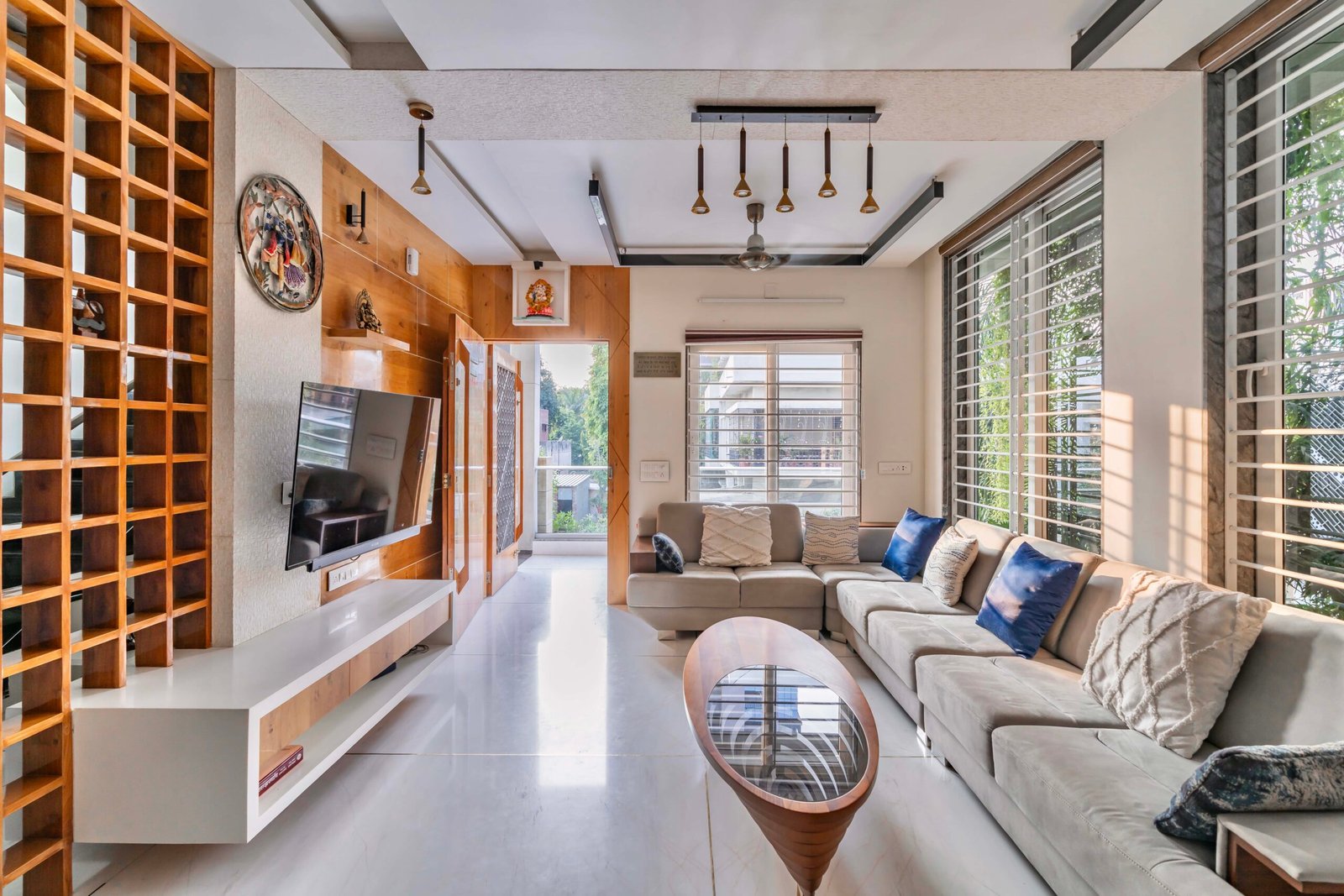 - Modern bungalow facade with exposed concrete, stone cladding, and wooden louvers in Ahmedabad - Sandstone-clad entrance with illuminated staircase and landscaped exterior - Open-plan living and dining area with geometric partition and modern chandelier - Master bedroom with blue textured wallpaper, wooden ceiling grid, and floating bedside units - Guest bedroom with geometric headboard, floating desk, and accent lighting - Polished flooring, natural wood finishes, and integrated lighting in modern residential interior