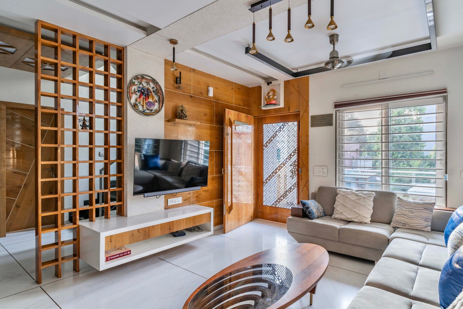 - Modern bungalow facade with exposed concrete, stone cladding, and wooden louvers in Ahmedabad - Sandstone-clad entrance with illuminated staircase and landscaped exterior - Open-plan living and dining area with geometric partition and modern chandelier - Master bedroom with blue textured wallpaper, wooden ceiling grid, and floating bedside units - Guest bedroom with geometric headboard, floating desk, and accent lighting - Polished flooring, natural wood finishes, and integrated lighting in modern residential interior