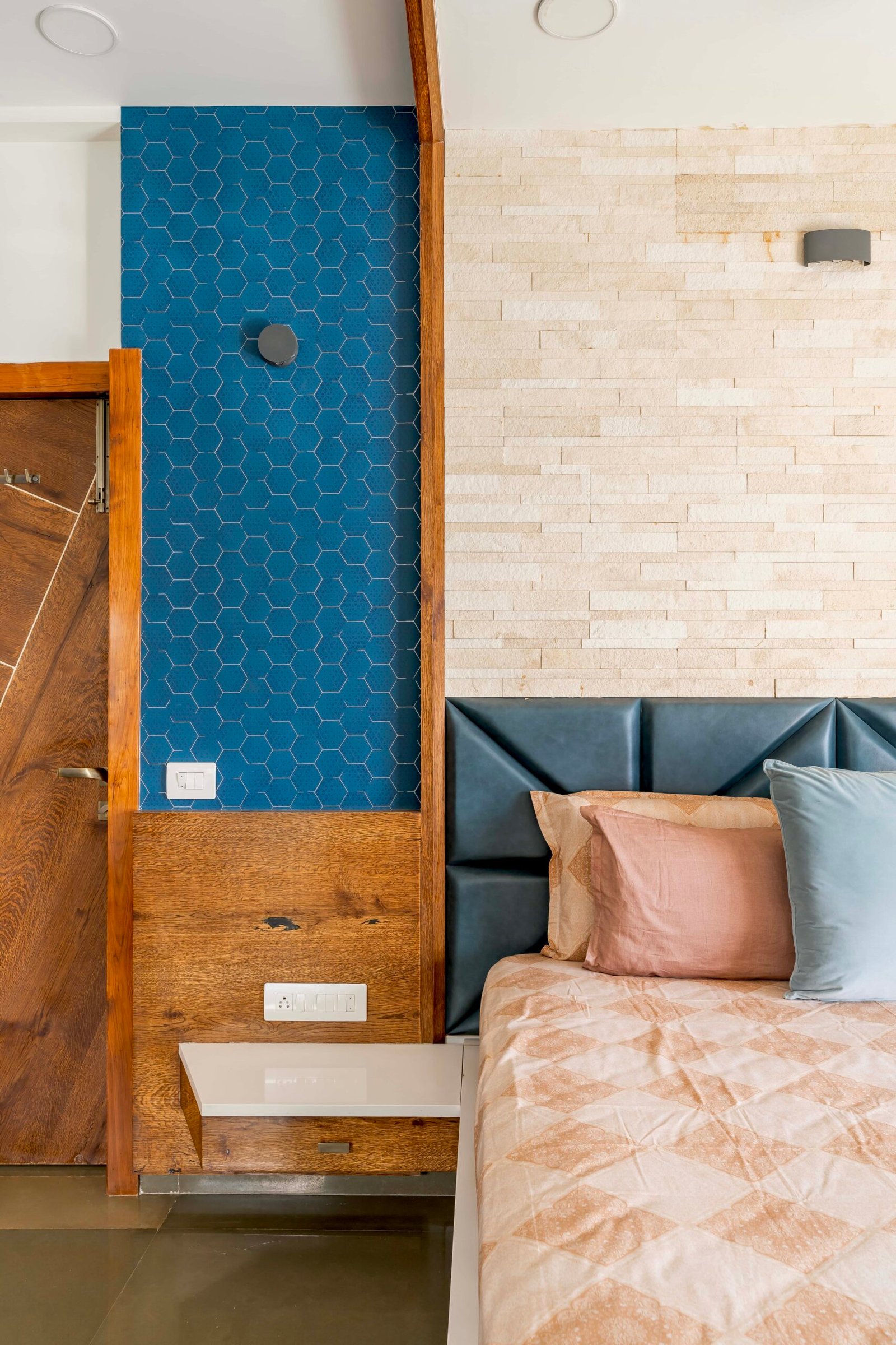 - Modern bungalow facade with exposed concrete, stone cladding, and wooden louvers in Ahmedabad - Sandstone-clad entrance with illuminated staircase and landscaped exterior - Open-plan living and dining area with geometric partition and modern chandelier - Master bedroom with blue textured wallpaper, wooden ceiling grid, and floating bedside units - Guest bedroom with geometric headboard, floating desk, and accent lighting - Polished flooring, natural wood finishes, and integrated lighting in modern residential interior
