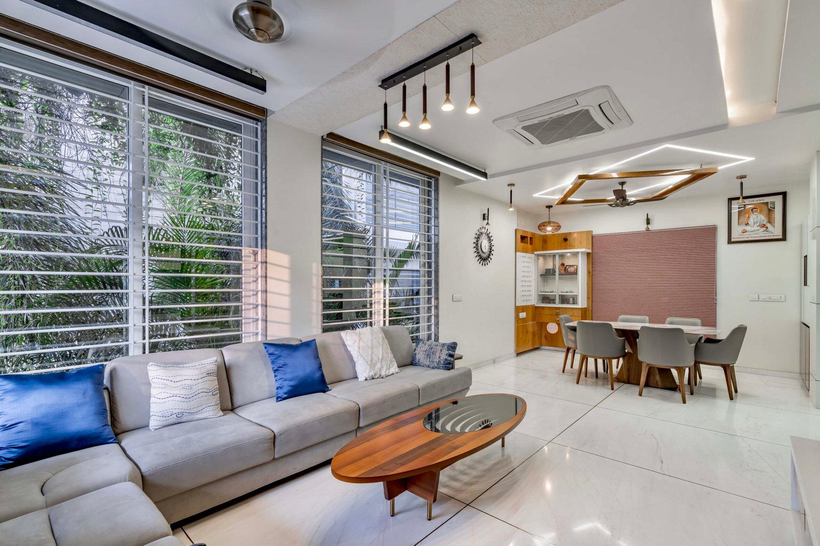 - Modern bungalow facade with exposed concrete, stone cladding, and wooden louvers in Ahmedabad - Sandstone-clad entrance with illuminated staircase and landscaped exterior - Open-plan living and dining area with geometric partition and modern chandelier - Master bedroom with blue textured wallpaper, wooden ceiling grid, and floating bedside units - Guest bedroom with geometric headboard, floating desk, and accent lighting - Polished flooring, natural wood finishes, and integrated lighting in modern residential interior