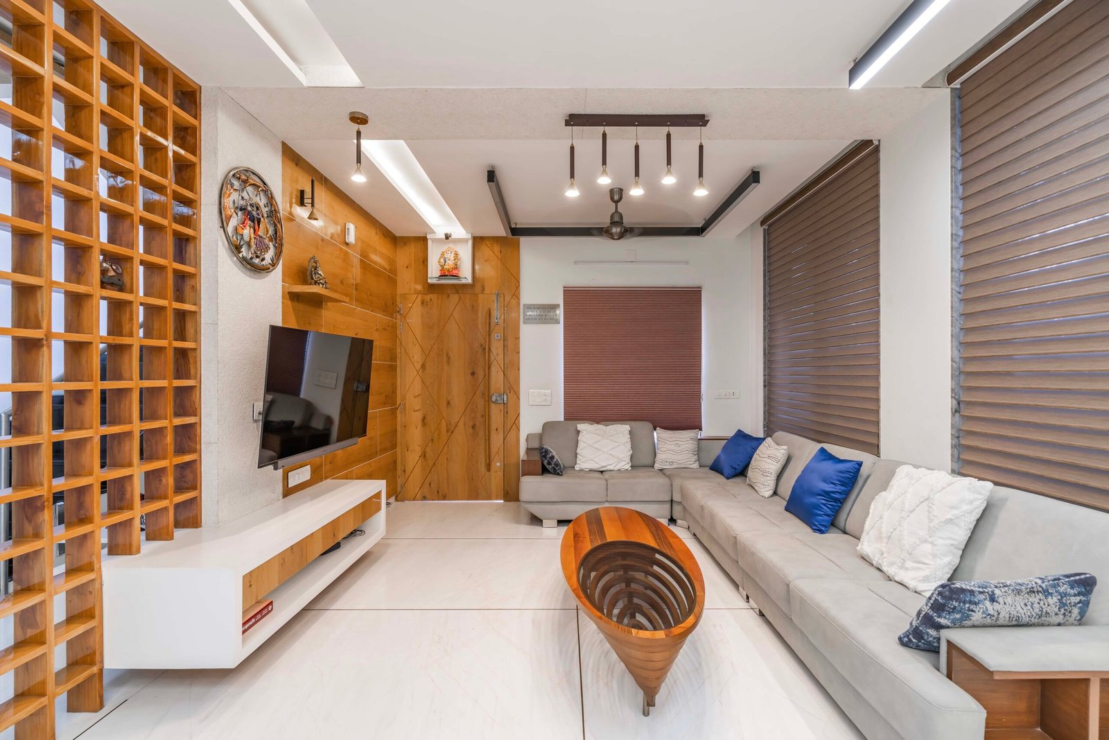 - Modern bungalow facade with exposed concrete, stone cladding, and wooden louvers in Ahmedabad - Sandstone-clad entrance with illuminated staircase and landscaped exterior - Open-plan living and dining area with geometric partition and modern chandelier - Master bedroom with blue textured wallpaper, wooden ceiling grid, and floating bedside units - Guest bedroom with geometric headboard, floating desk, and accent lighting - Polished flooring, natural wood finishes, and integrated lighting in modern residential interior