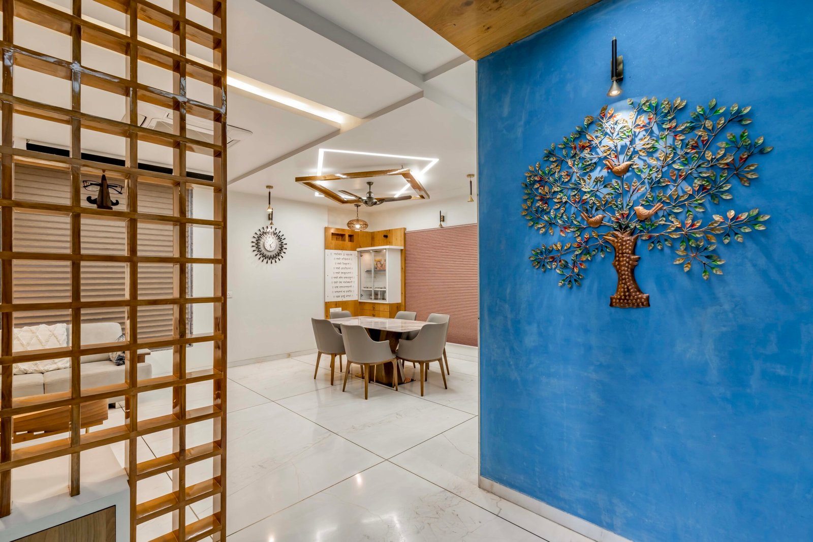 - Modern bungalow facade with exposed concrete, stone cladding, and wooden louvers in Ahmedabad - Sandstone-clad entrance with illuminated staircase and landscaped exterior - Open-plan living and dining area with geometric partition and modern chandelier - Master bedroom with blue textured wallpaper, wooden ceiling grid, and floating bedside units - Guest bedroom with geometric headboard, floating desk, and accent lighting - Polished flooring, natural wood finishes, and integrated lighting in modern residential interior