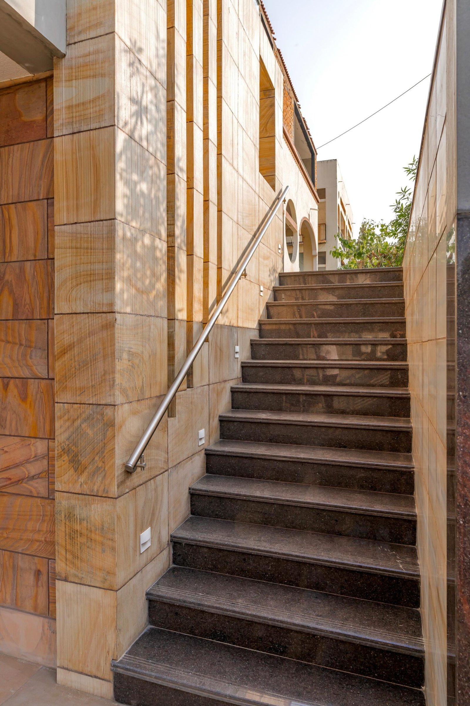 - Modern bungalow facade with exposed concrete, stone cladding, and wooden louvers in Ahmedabad - Sandstone-clad entrance with illuminated staircase and landscaped exterior - Open-plan living and dining area with geometric partition and modern chandelier - Master bedroom with blue textured wallpaper, wooden ceiling grid, and floating bedside units - Guest bedroom with geometric headboard, floating desk, and accent lighting - Polished flooring, natural wood finishes, and integrated lighting in modern residential interior