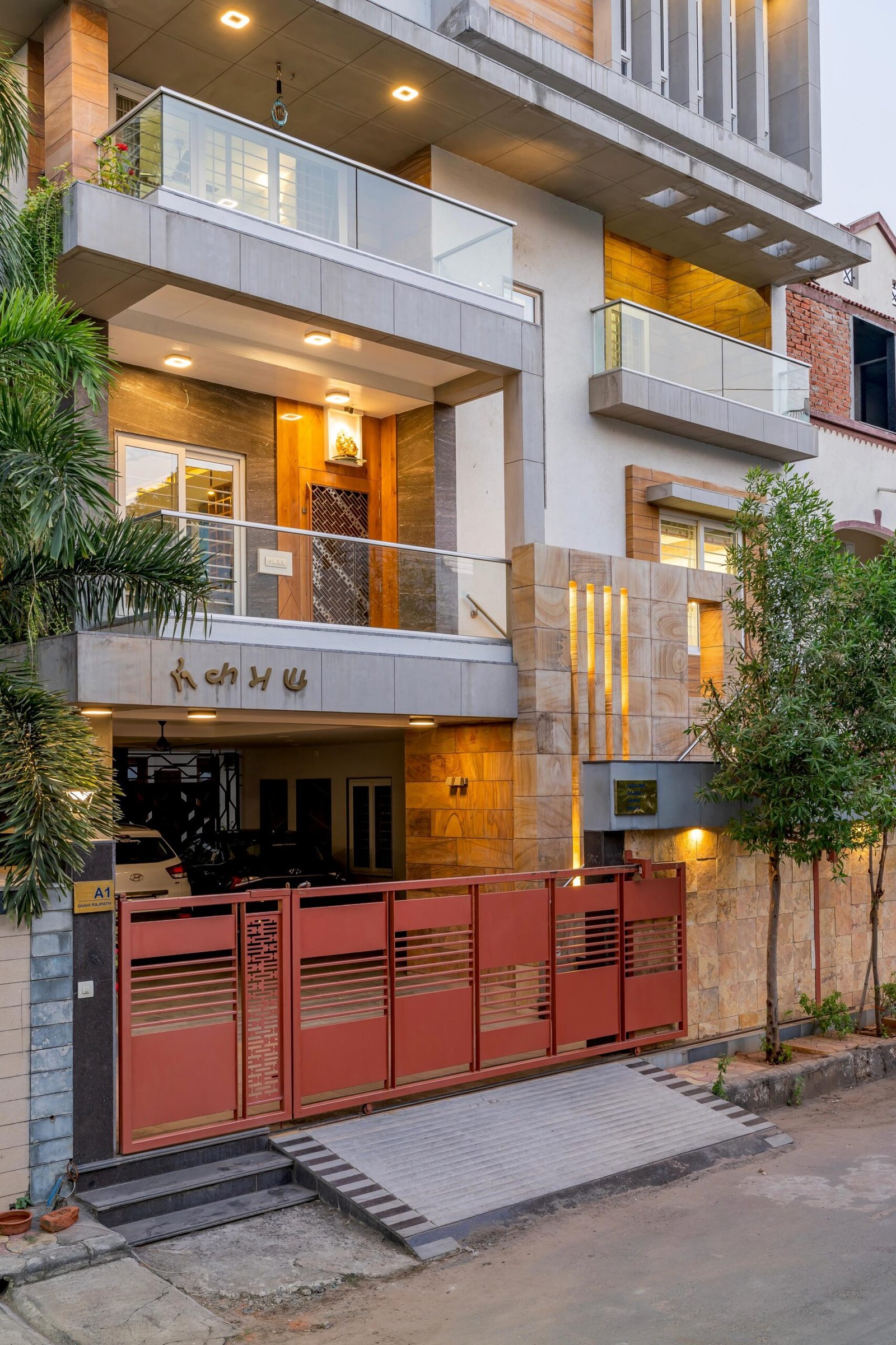 - Modern bungalow facade with exposed concrete, stone cladding, and wooden louvers in Ahmedabad - Sandstone-clad entrance with illuminated staircase and landscaped exterior - Open-plan living and dining area with geometric partition and modern chandelier - Master bedroom with blue textured wallpaper, wooden ceiling grid, and floating bedside units - Guest bedroom with geometric headboard, floating desk, and accent lighting - Polished flooring, natural wood finishes, and integrated lighting in modern residential interior