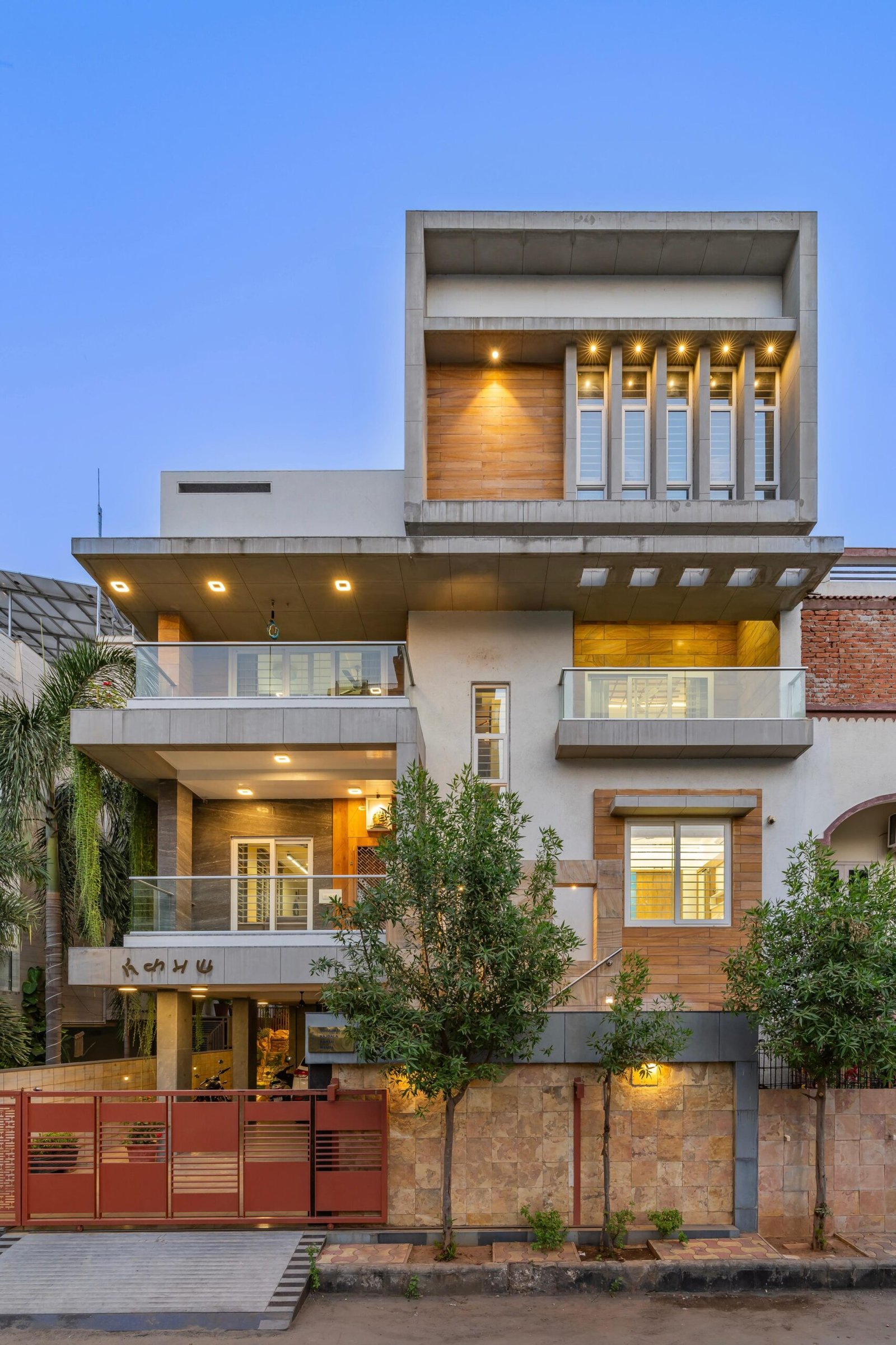 - Modern bungalow facade with exposed concrete, stone cladding, and wooden louvers in Ahmedabad - Sandstone-clad entrance with illuminated staircase and landscaped exterior - Open-plan living and dining area with geometric partition and modern chandelier - Master bedroom with blue textured wallpaper, wooden ceiling grid, and floating bedside units - Guest bedroom with geometric headboard, floating desk, and accent lighting - Polished flooring, natural wood finishes, and integrated lighting in modern residential interior