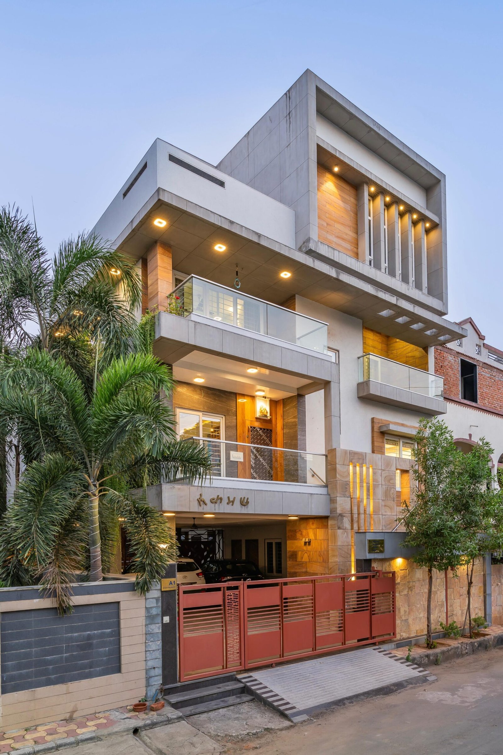 - Modern bungalow facade with exposed concrete, stone cladding, and wooden louvers in Ahmedabad - Sandstone-clad entrance with illuminated staircase and landscaped exterior - Open-plan living and dining area with geometric partition and modern chandelier - Master bedroom with blue textured wallpaper, wooden ceiling grid, and floating bedside units - Guest bedroom with geometric headboard, floating desk, and accent lighting - Polished flooring, natural wood finishes, and integrated lighting in modern residential interior