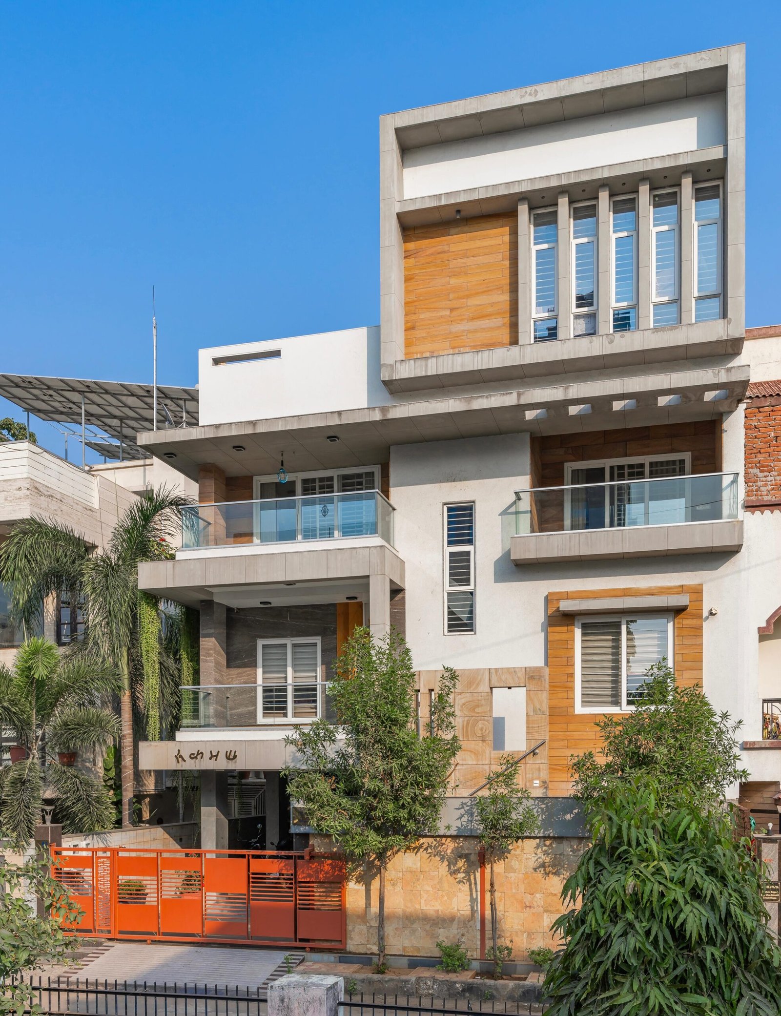 - Modern bungalow facade with exposed concrete, stone cladding, and wooden louvers in Ahmedabad - Sandstone-clad entrance with illuminated staircase and landscaped exterior - Open-plan living and dining area with geometric partition and modern chandelier - Master bedroom with blue textured wallpaper, wooden ceiling grid, and floating bedside units - Guest bedroom with geometric headboard, floating desk, and accent lighting - Polished flooring, natural wood finishes, and integrated lighting in modern residential interior