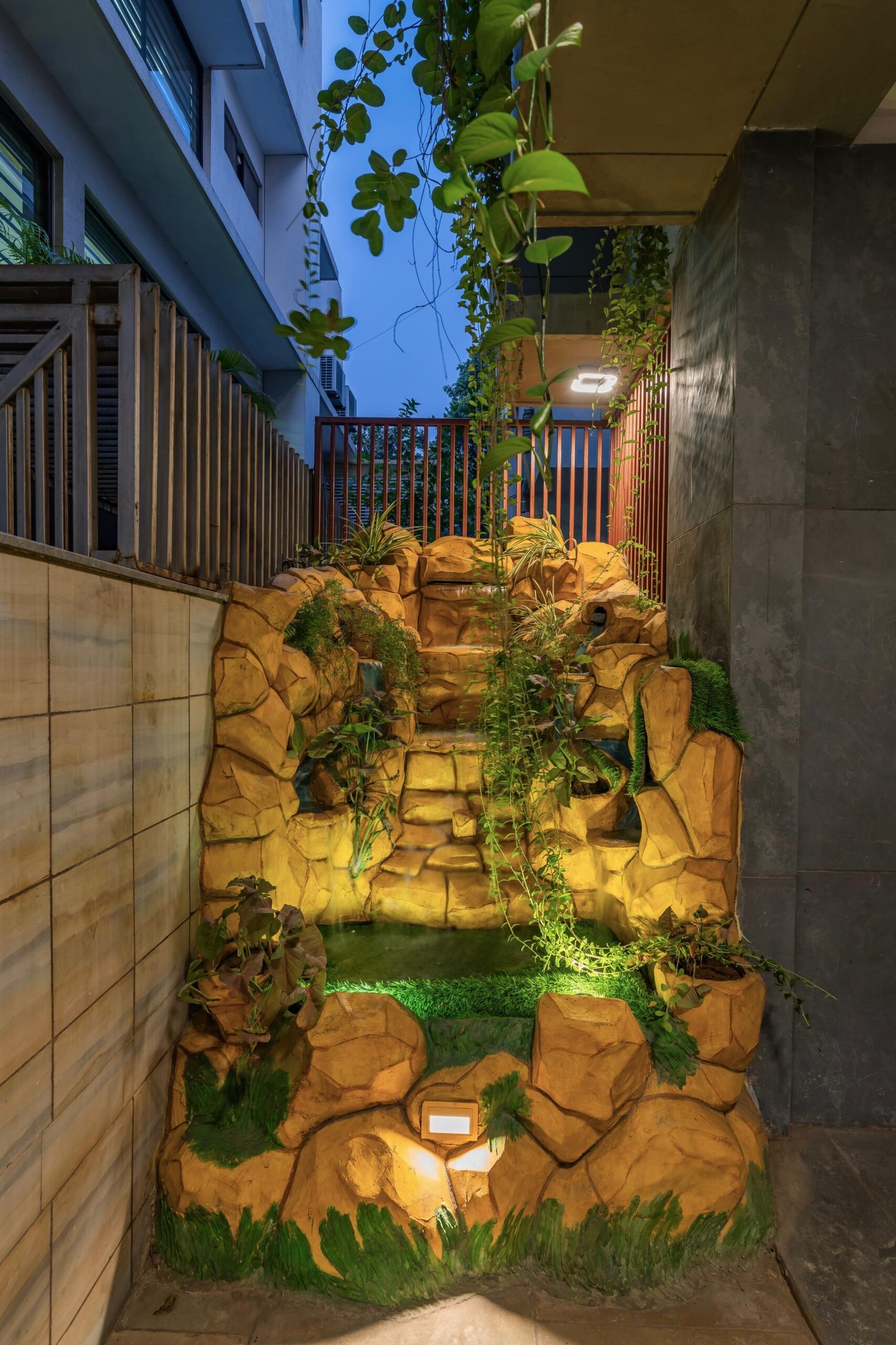 - Modern bungalow facade with exposed concrete, stone cladding, and wooden louvers in Ahmedabad - Sandstone-clad entrance with illuminated staircase and landscaped exterior - Open-plan living and dining area with geometric partition and modern chandelier - Master bedroom with blue textured wallpaper, wooden ceiling grid, and floating bedside units - Guest bedroom with geometric headboard, floating desk, and accent lighting - Polished flooring, natural wood finishes, and integrated lighting in modern residential interior