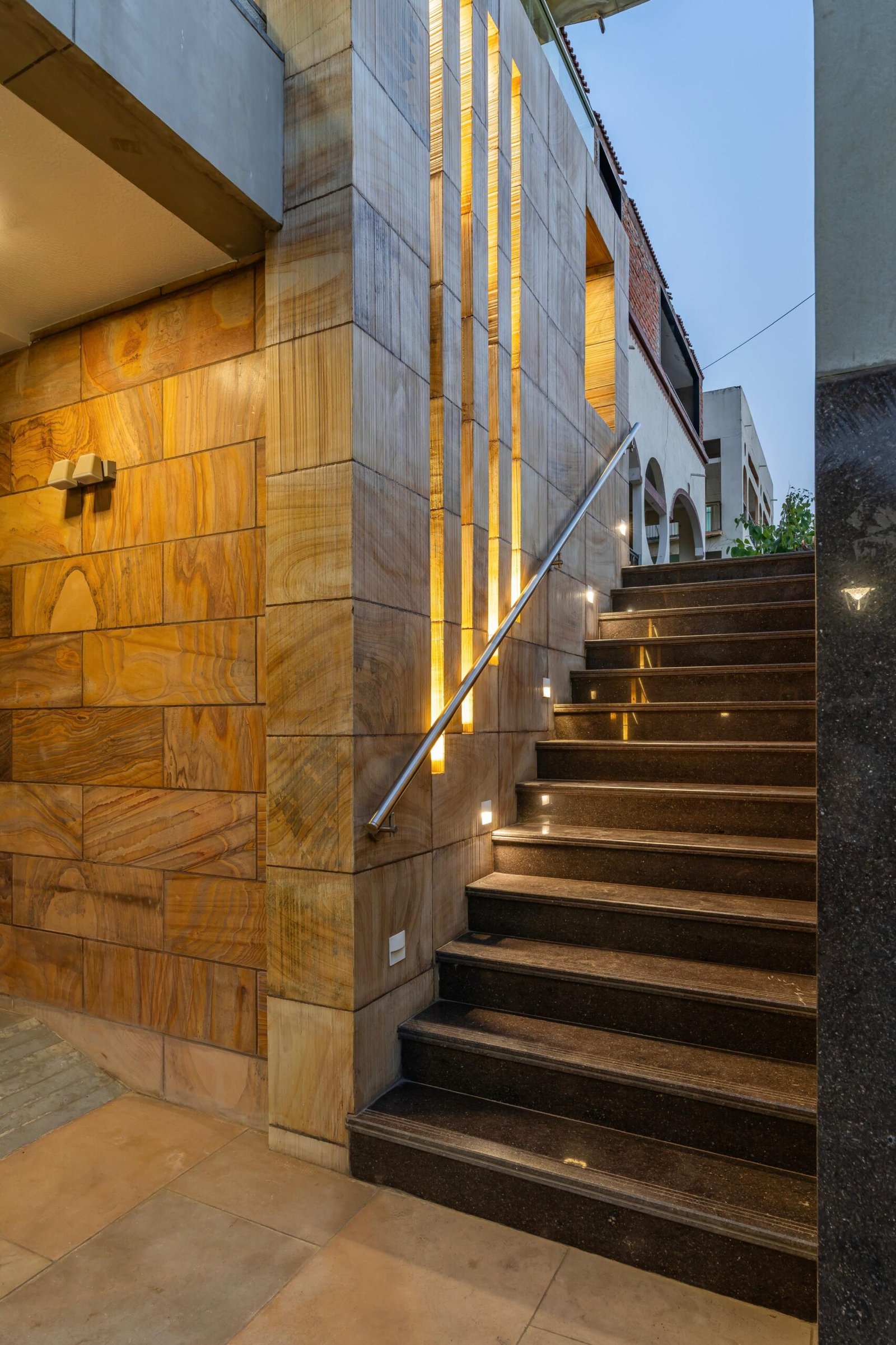 - Modern bungalow facade with exposed concrete, stone cladding, and wooden louvers in Ahmedabad - Sandstone-clad entrance with illuminated staircase and landscaped exterior - Open-plan living and dining area with geometric partition and modern chandelier - Master bedroom with blue textured wallpaper, wooden ceiling grid, and floating bedside units - Guest bedroom with geometric headboard, floating desk, and accent lighting - Polished flooring, natural wood finishes, and integrated lighting in modern residential interior