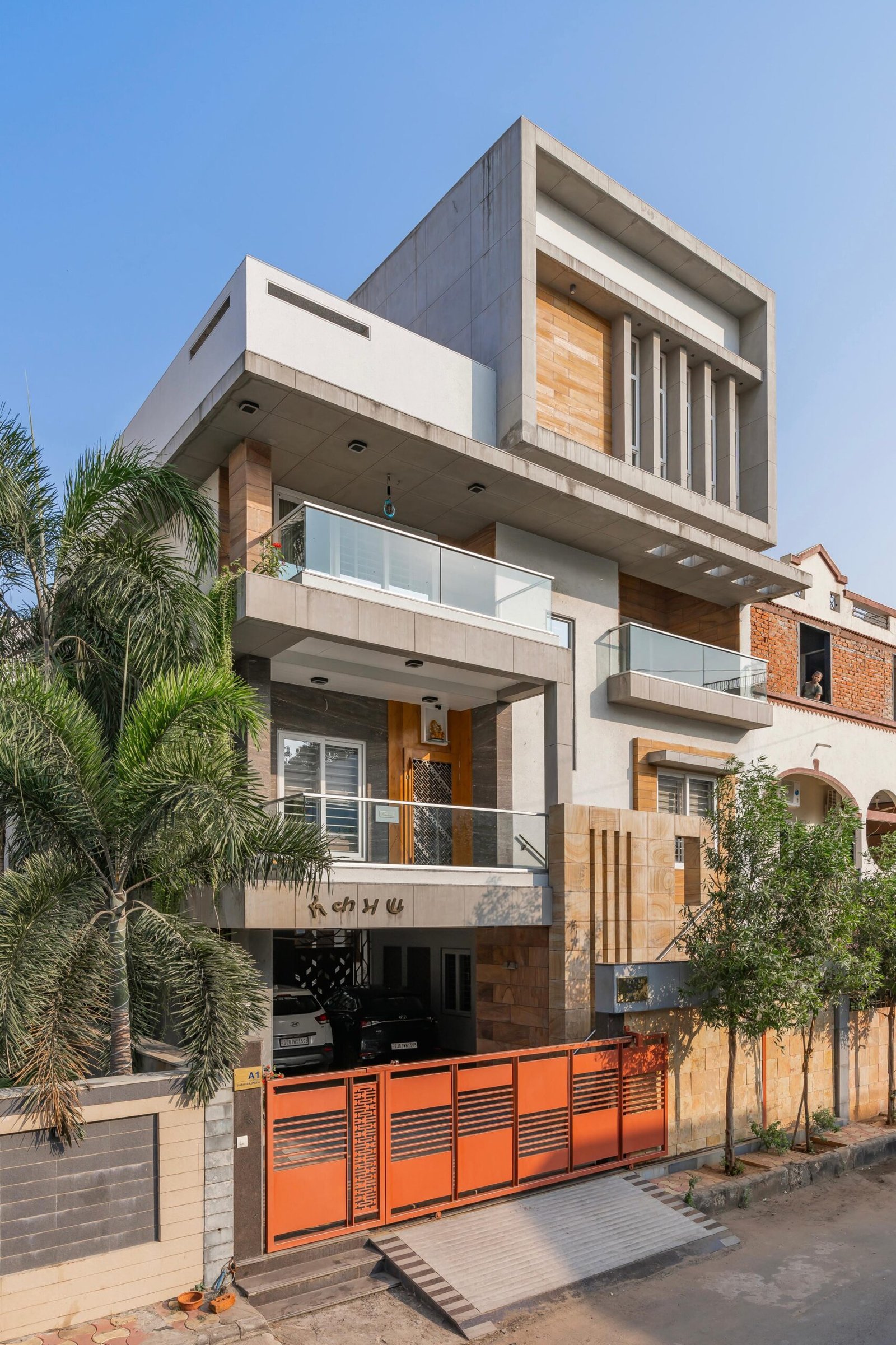 - Modern bungalow facade with exposed concrete, stone cladding, and wooden louvers in Ahmedabad - Sandstone-clad entrance with illuminated staircase and landscaped exterior - Open-plan living and dining area with geometric partition and modern chandelier - Master bedroom with blue textured wallpaper, wooden ceiling grid, and floating bedside units - Guest bedroom with geometric headboard, floating desk, and accent lighting - Polished flooring, natural wood finishes, and integrated lighting in modern residential interior