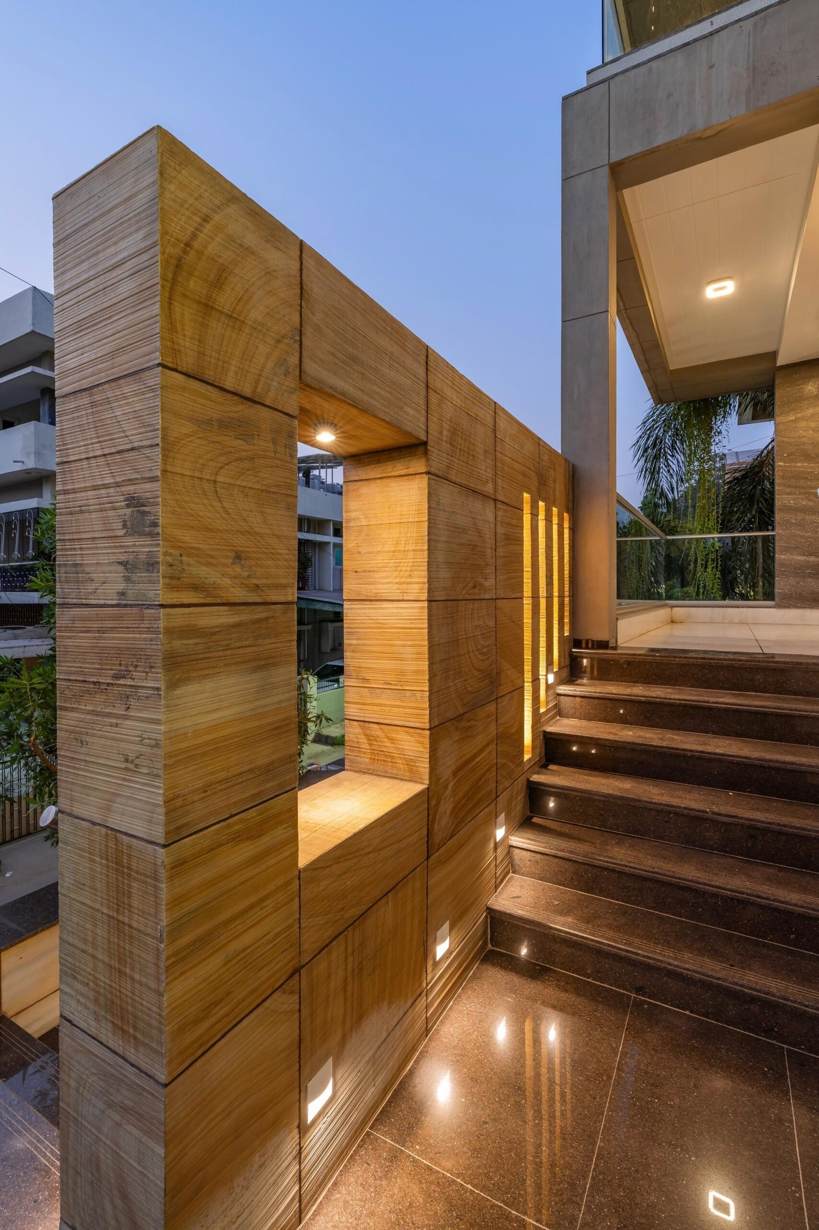 - Modern bungalow facade with exposed concrete, stone cladding, and wooden louvers in Ahmedabad - Sandstone-clad entrance with illuminated staircase and landscaped exterior - Open-plan living and dining area with geometric partition and modern chandelier - Master bedroom with blue textured wallpaper, wooden ceiling grid, and floating bedside units - Guest bedroom with geometric headboard, floating desk, and accent lighting - Polished flooring, natural wood finishes, and integrated lighting in modern residential interior
