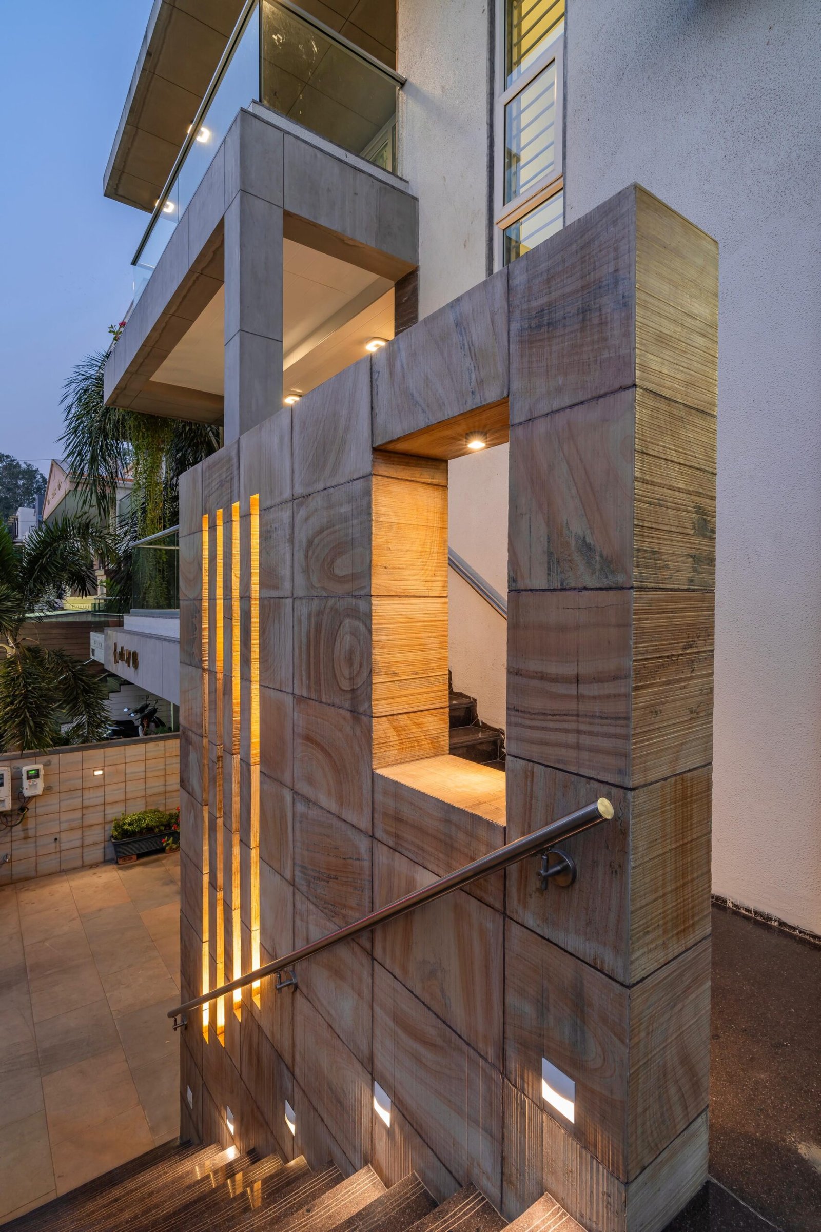 - Modern bungalow facade with exposed concrete, stone cladding, and wooden louvers in Ahmedabad - Sandstone-clad entrance with illuminated staircase and landscaped exterior - Open-plan living and dining area with geometric partition and modern chandelier - Master bedroom with blue textured wallpaper, wooden ceiling grid, and floating bedside units - Guest bedroom with geometric headboard, floating desk, and accent lighting - Polished flooring, natural wood finishes, and integrated lighting in modern residential interior