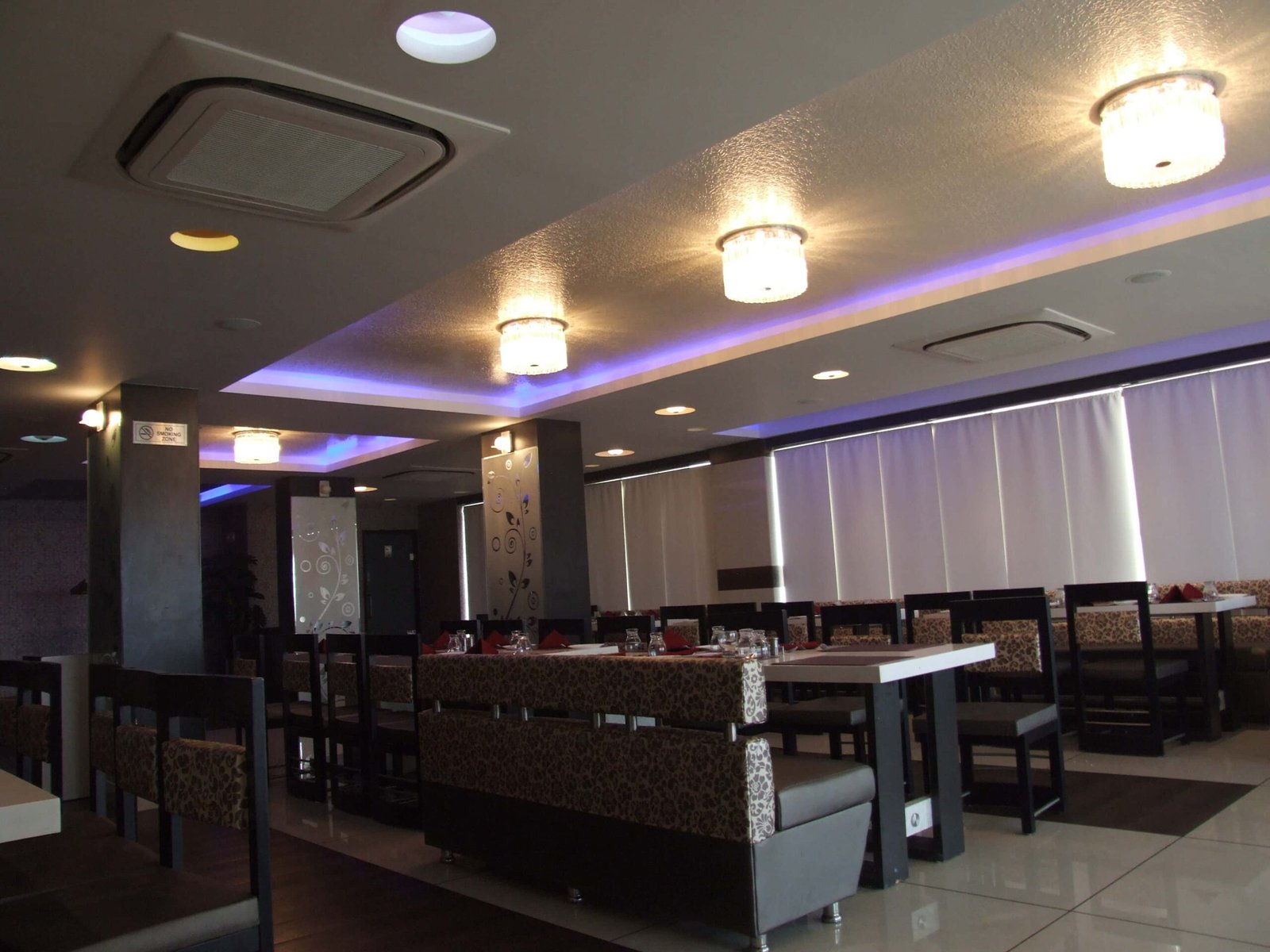 - Foyer of Shabri Restaurant & Banquet Hall with bold geometric wall art and ambient lighting - Dining area featuring plush banquet seating arranged around contemporary chandeliers - Intimate private dining corner with upholstered booths and dramatic accent walls - Grand banquet hall set for an event with long tables, floral centerpieces, and ambient uplighting - Artistic mural and bespoke furniture framing the restaurant’s main corridor