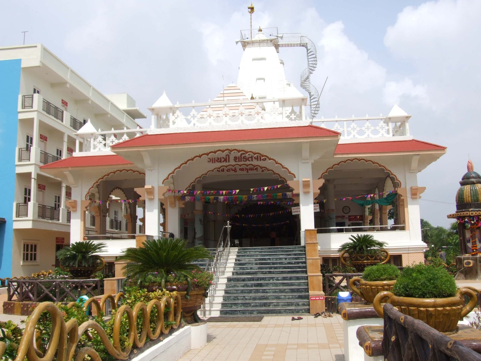 - Front view of the Gayatri Shaktipith Temple showcasing Himalayan-inspired shivalay architecture against a clear sky - Ferrocement artificial wood beams lining a landscaped pathway beside reflecting pools - Aerial perspective of the temple campus with serene gardens, pathways, and mountain-style temple roofs - Devotees walking through manicured gardens flanked by ferrocement sculptures resembling timber structures - Close-up of the shivalay temple’s ornate façade with traditional carvings and eco-centric design elements