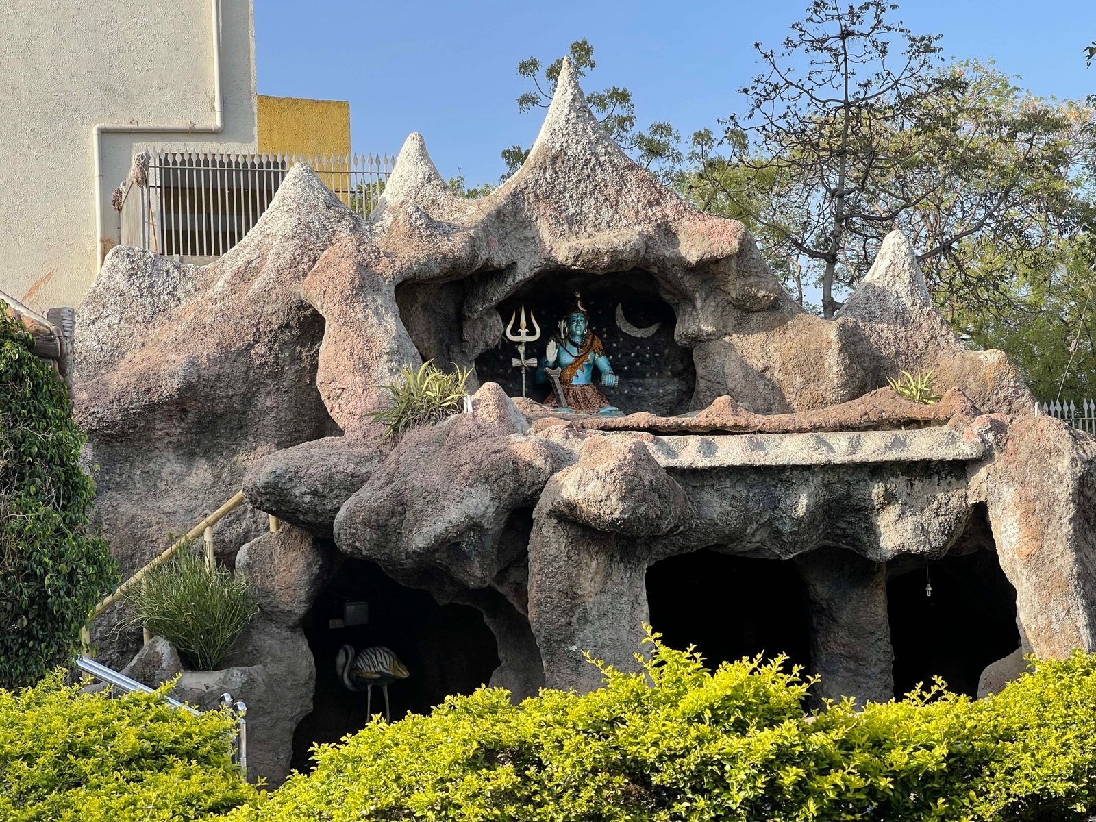 - Front view of the Gayatri Shaktipith Temple showcasing Himalayan-inspired shivalay architecture against a clear sky - Ferrocement artificial wood beams lining a landscaped pathway beside reflecting pools - Aerial perspective of the temple campus with serene gardens, pathways, and mountain-style temple roofs - Devotees walking through manicured gardens flanked by ferrocement sculptures resembling timber structures - Close-up of the shivalay temple’s ornate façade with traditional carvings and eco-centric design elements
