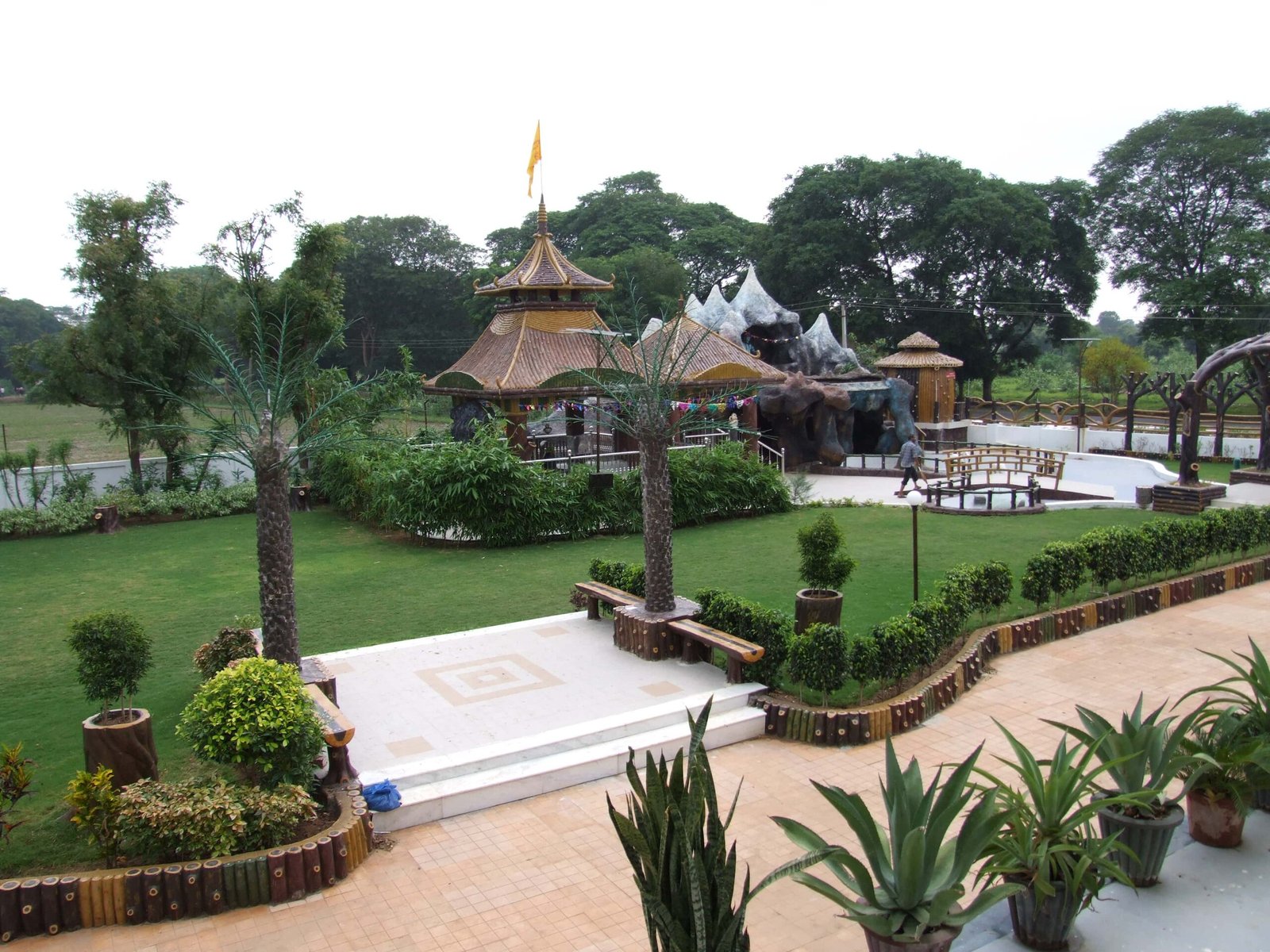 - Front view of the Gayatri Shaktipith Temple showcasing Himalayan-inspired shivalay architecture against a clear sky - Ferrocement artificial wood beams lining a landscaped pathway beside reflecting pools - Aerial perspective of the temple campus with serene gardens, pathways, and mountain-style temple roofs - Devotees walking through manicured gardens flanked by ferrocement sculptures resembling timber structures - Close-up of the shivalay temple’s ornate façade with traditional carvings and eco-centric design elements