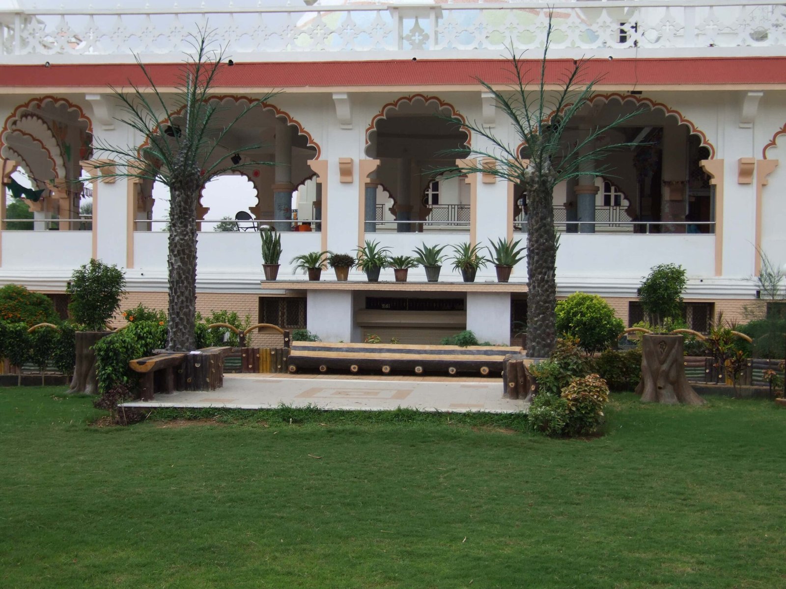 - Front view of the Gayatri Shaktipith Temple showcasing Himalayan-inspired shivalay architecture against a clear sky - Ferrocement artificial wood beams lining a landscaped pathway beside reflecting pools - Aerial perspective of the temple campus with serene gardens, pathways, and mountain-style temple roofs - Devotees walking through manicured gardens flanked by ferrocement sculptures resembling timber structures - Close-up of the shivalay temple’s ornate façade with traditional carvings and eco-centric design elements