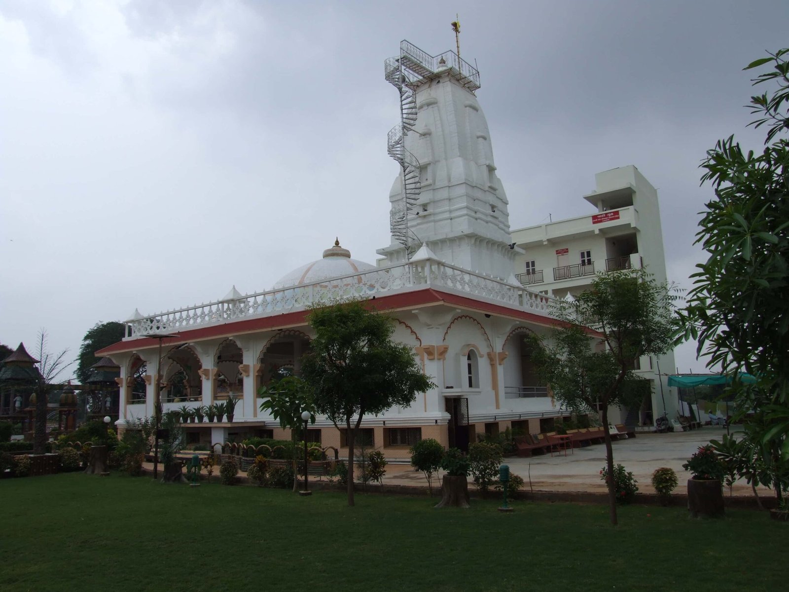 - Front view of the Gayatri Shaktipith Temple showcasing Himalayan-inspired shivalay architecture against a clear sky - Ferrocement artificial wood beams lining a landscaped pathway beside reflecting pools - Aerial perspective of the temple campus with serene gardens, pathways, and mountain-style temple roofs - Devotees walking through manicured gardens flanked by ferrocement sculptures resembling timber structures - Close-up of the shivalay temple’s ornate façade with traditional carvings and eco-centric design elements