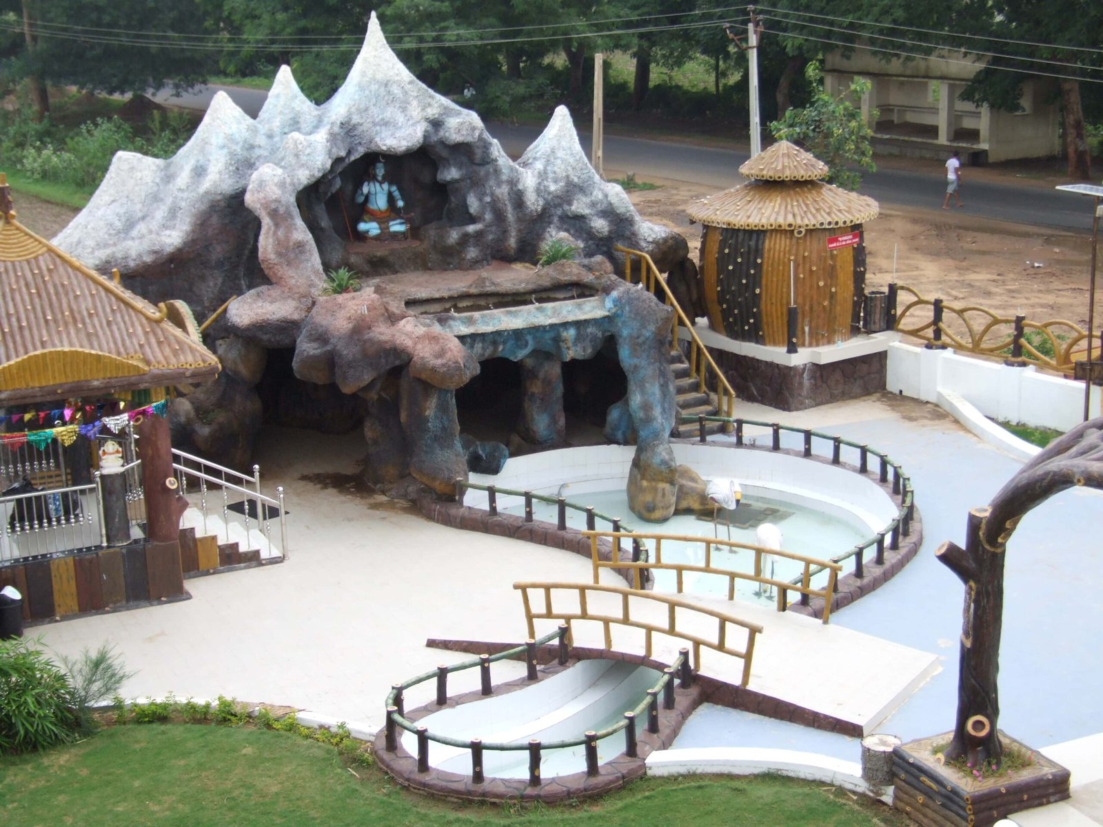 - Front view of the Gayatri Shaktipith Temple showcasing Himalayan-inspired shivalay architecture against a clear sky - Ferrocement artificial wood beams lining a landscaped pathway beside reflecting pools - Aerial perspective of the temple campus with serene gardens, pathways, and mountain-style temple roofs - Devotees walking through manicured gardens flanked by ferrocement sculptures resembling timber structures - Close-up of the shivalay temple’s ornate façade with traditional carvings and eco-centric design elements