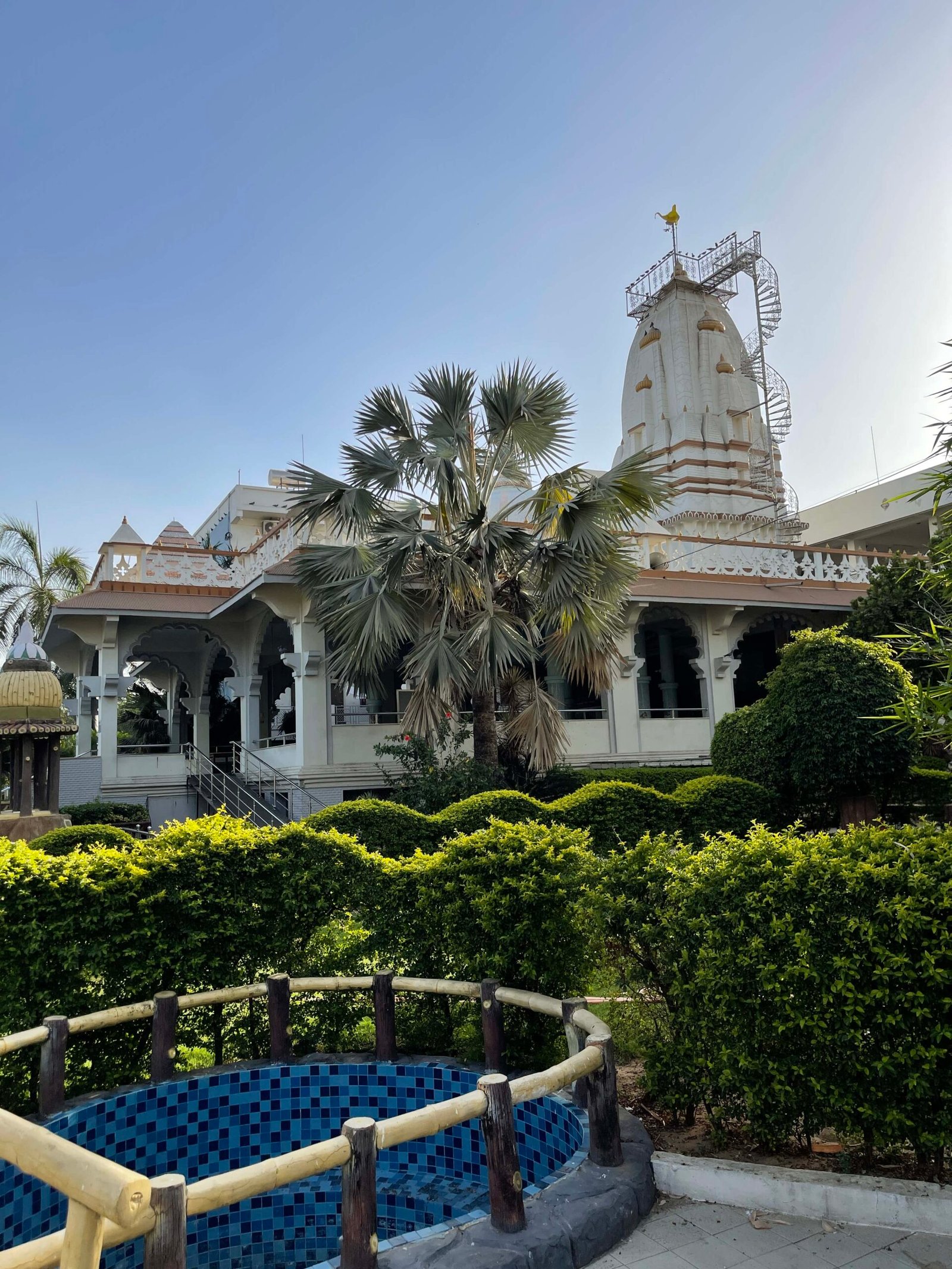 - Front view of the Gayatri Shaktipith Temple showcasing Himalayan-inspired shivalay architecture against a clear sky - Ferrocement artificial wood beams lining a landscaped pathway beside reflecting pools - Aerial perspective of the temple campus with serene gardens, pathways, and mountain-style temple roofs - Devotees walking through manicured gardens flanked by ferrocement sculptures resembling timber structures - Close-up of the shivalay temple’s ornate façade with traditional carvings and eco-centric design elements