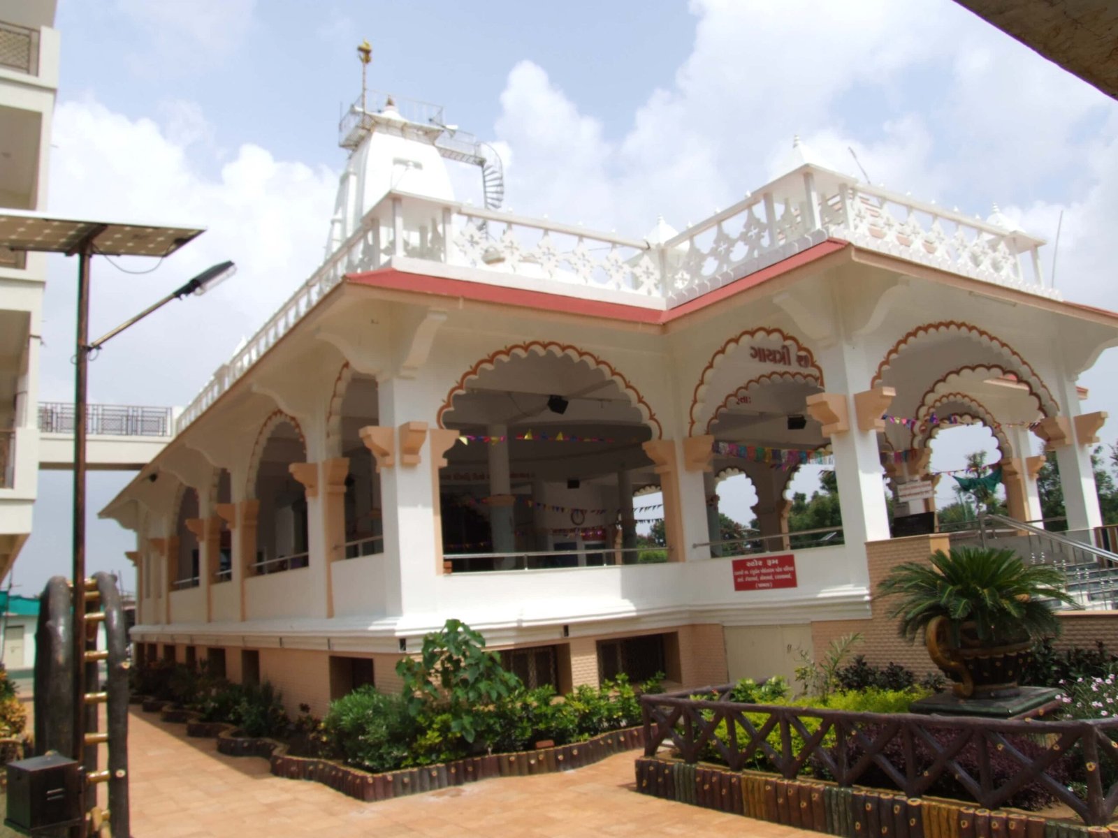 - Front view of the Gayatri Shaktipith Temple showcasing Himalayan-inspired shivalay architecture against a clear sky - Ferrocement artificial wood beams lining a landscaped pathway beside reflecting pools - Aerial perspective of the temple campus with serene gardens, pathways, and mountain-style temple roofs - Devotees walking through manicured gardens flanked by ferrocement sculptures resembling timber structures - Close-up of the shivalay temple’s ornate façade with traditional carvings and eco-centric design elements
