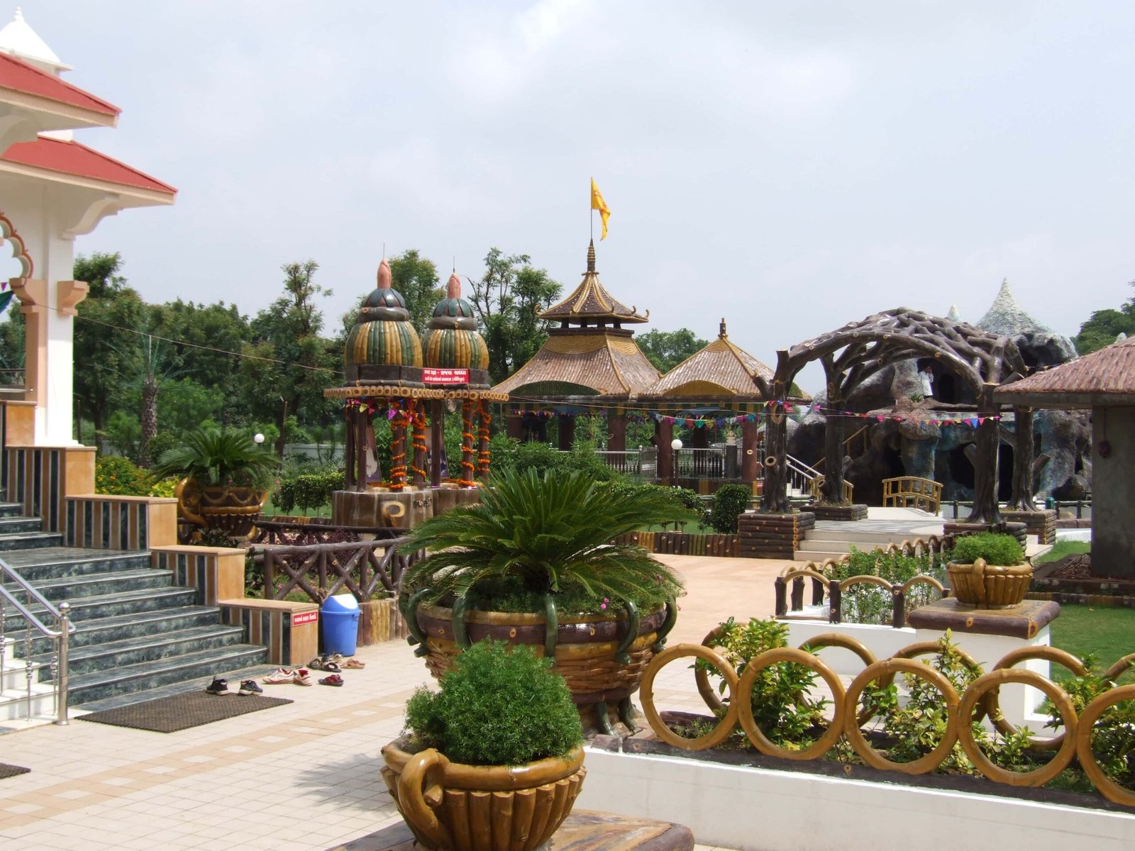 - Front view of the Gayatri Shaktipith Temple showcasing Himalayan-inspired shivalay architecture against a clear sky - Ferrocement artificial wood beams lining a landscaped pathway beside reflecting pools - Aerial perspective of the temple campus with serene gardens, pathways, and mountain-style temple roofs - Devotees walking through manicured gardens flanked by ferrocement sculptures resembling timber structures - Close-up of the shivalay temple’s ornate façade with traditional carvings and eco-centric design elements