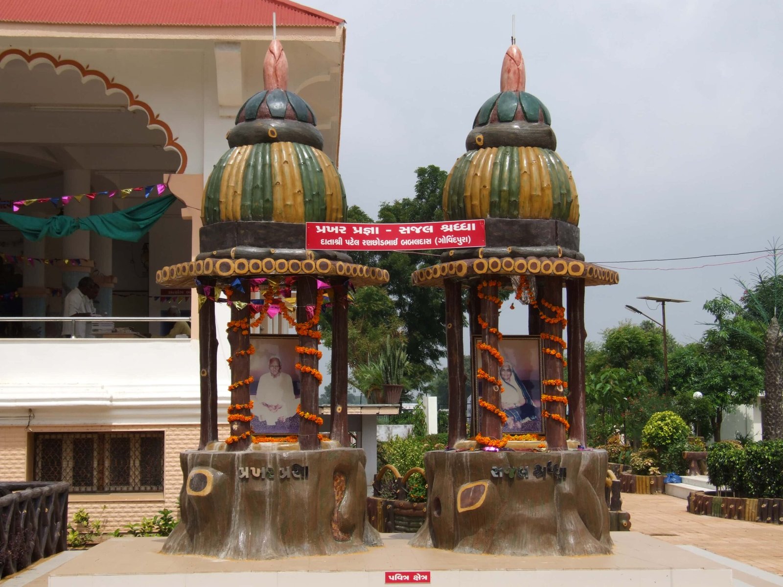 - Front view of the Gayatri Shaktipith Temple showcasing Himalayan-inspired shivalay architecture against a clear sky - Ferrocement artificial wood beams lining a landscaped pathway beside reflecting pools - Aerial perspective of the temple campus with serene gardens, pathways, and mountain-style temple roofs - Devotees walking through manicured gardens flanked by ferrocement sculptures resembling timber structures - Close-up of the shivalay temple’s ornate façade with traditional carvings and eco-centric design elements