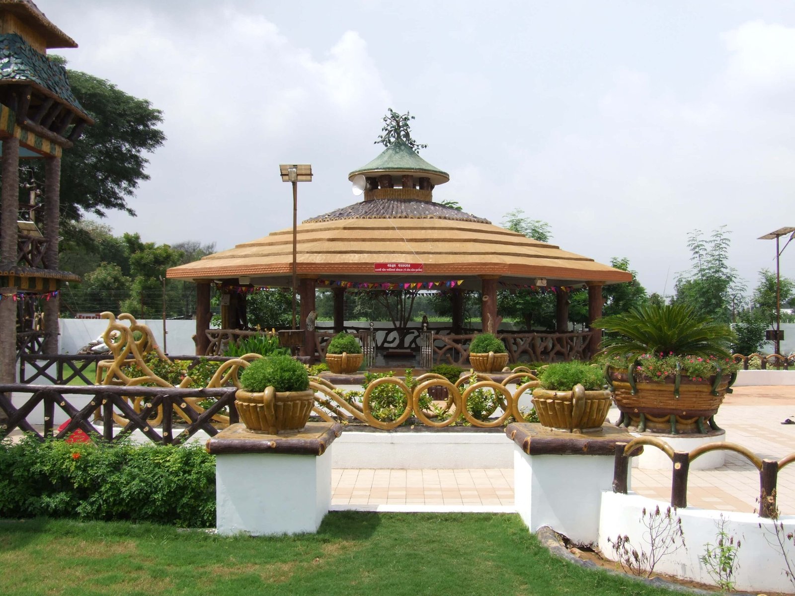 - Front view of the Gayatri Shaktipith Temple showcasing Himalayan-inspired shivalay architecture against a clear sky - Ferrocement artificial wood beams lining a landscaped pathway beside reflecting pools - Aerial perspective of the temple campus with serene gardens, pathways, and mountain-style temple roofs - Devotees walking through manicured gardens flanked by ferrocement sculptures resembling timber structures - Close-up of the shivalay temple’s ornate façade with traditional carvings and eco-centric design elements
