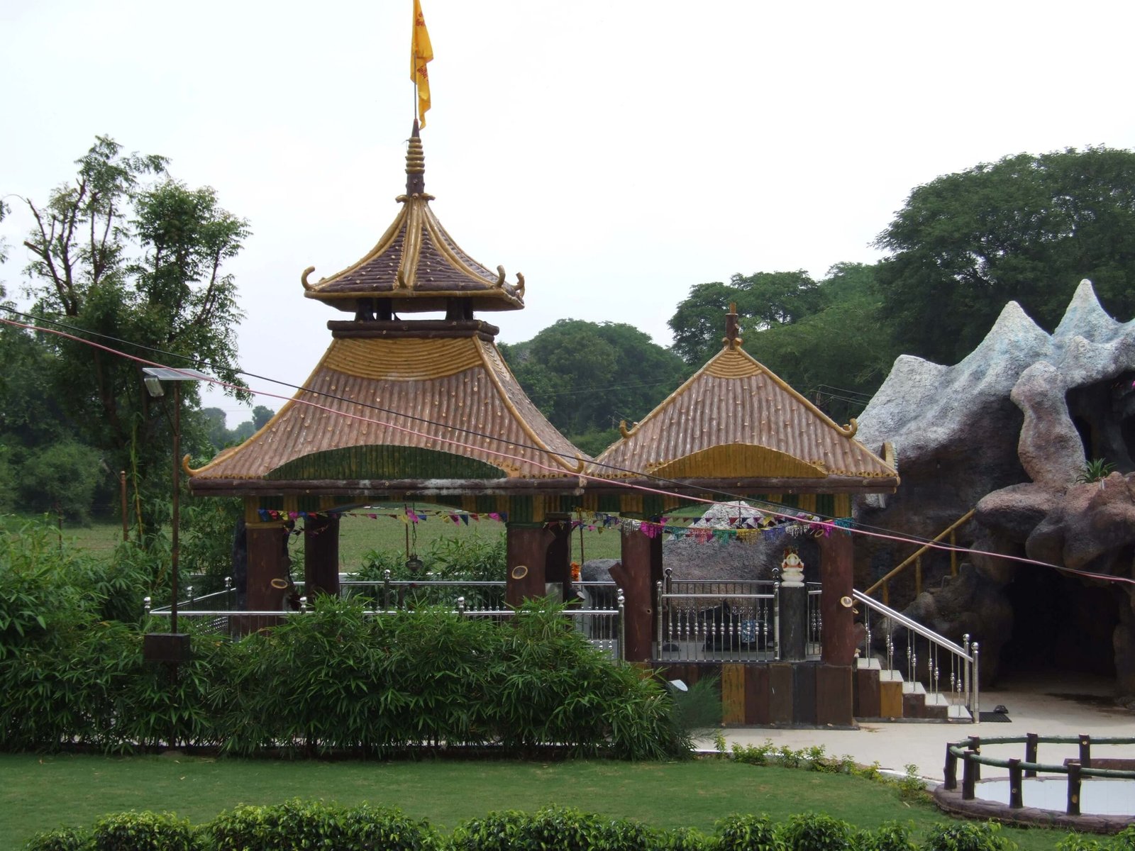 - Front view of the Gayatri Shaktipith Temple showcasing Himalayan-inspired shivalay architecture against a clear sky - Ferrocement artificial wood beams lining a landscaped pathway beside reflecting pools - Aerial perspective of the temple campus with serene gardens, pathways, and mountain-style temple roofs - Devotees walking through manicured gardens flanked by ferrocement sculptures resembling timber structures - Close-up of the shivalay temple’s ornate façade with traditional carvings and eco-centric design elements